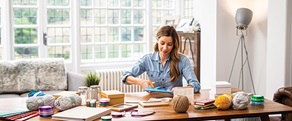 Une femme utilisant des ciseaux est assise à une table couverte de boîtes, de bocaux, de pelotes de laine et de dossiers.